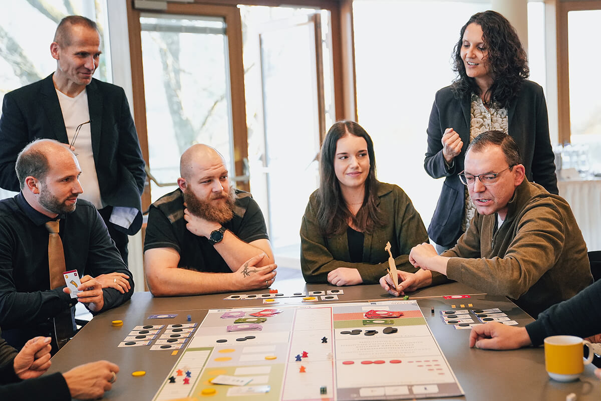 Mehrere Teilnehmende sitzen und stehen um einen Tisch und spielen Celsius - Die 2 Grad Challenge