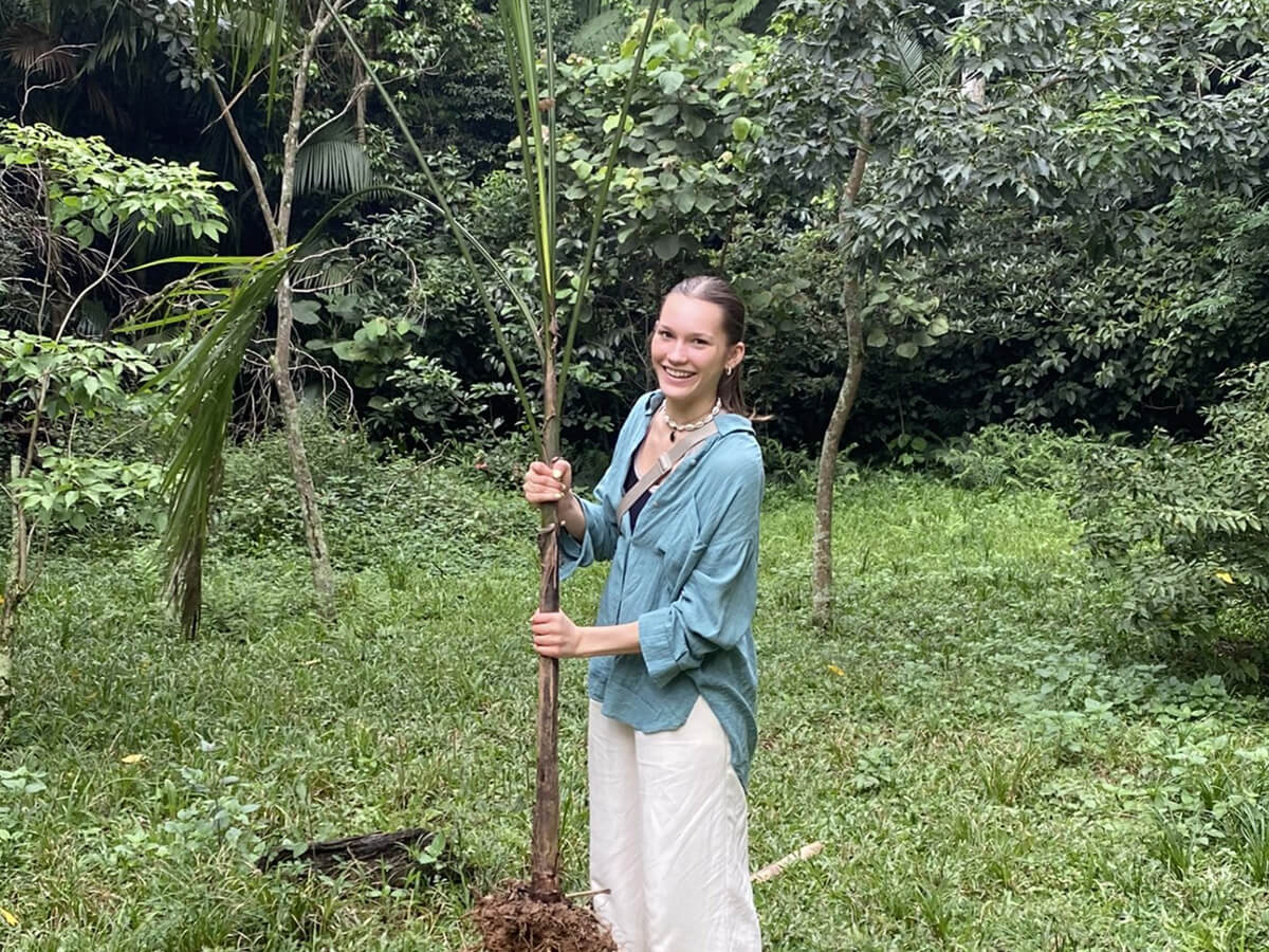 Lorena steht im Regenwald und hält eine junge Palme mit Wurzelballen in den Händen