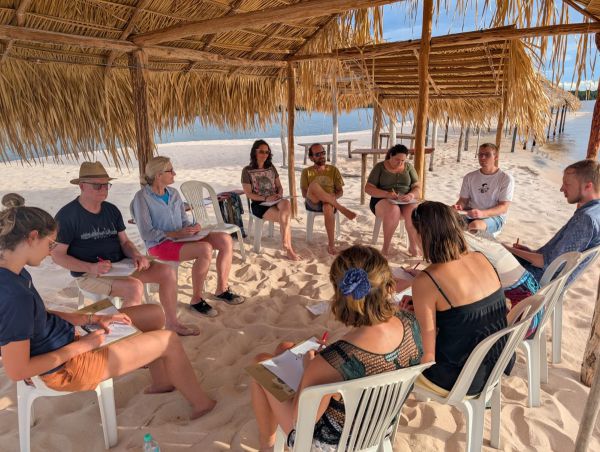 Mehrere Personen sitzen unter einem Palmdach am Strand und nehmen an einem Workshop teil