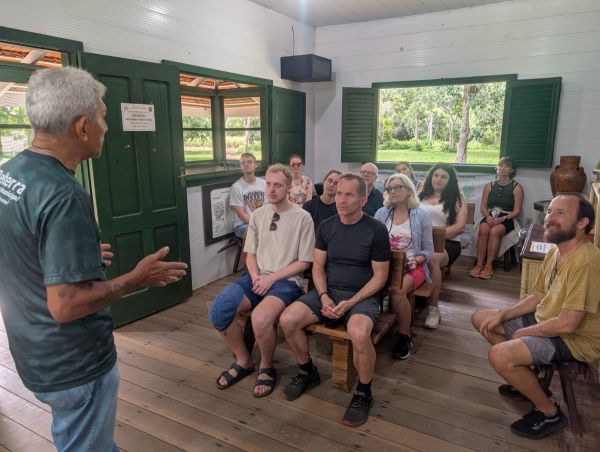 Eine Gruppe hört in einem offenen Holzhaus in Brasilien einer Frau zu, die vor einer Tafel spricht