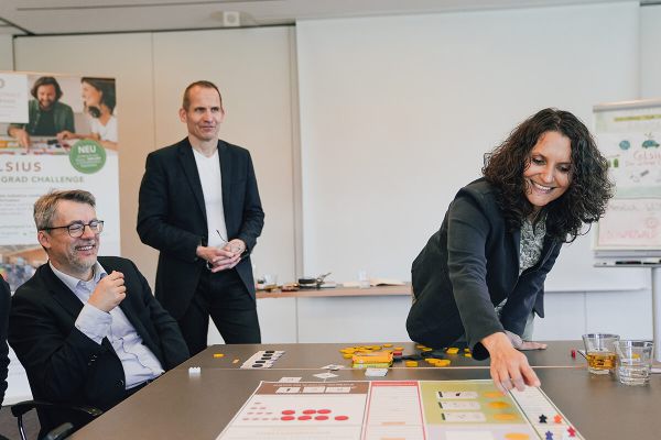 Eine Teilnehmerin legt Spielsteine auf das Spielfeld, während zwei Workshopleitende und ein weiterer Teilnehmer zusehen