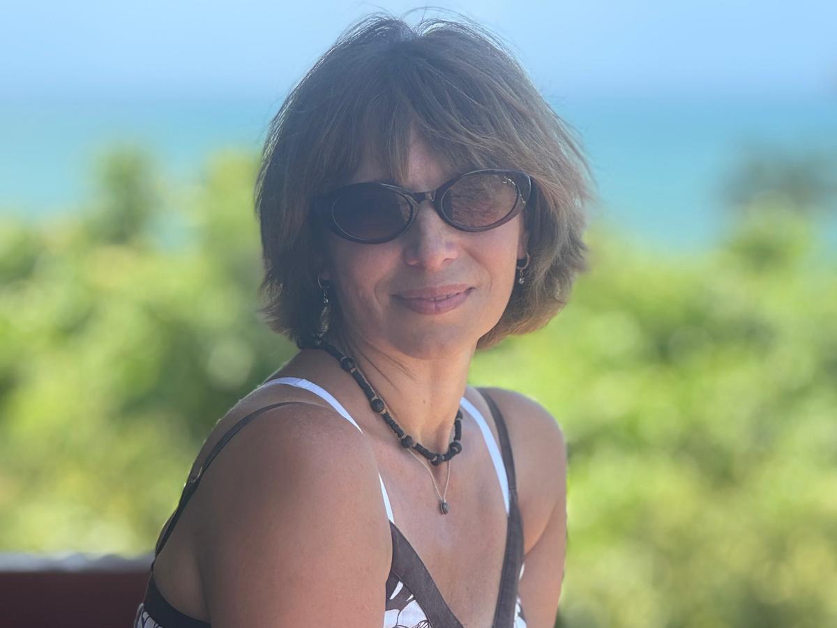 Portrait von Elena auf einer Holzterrasse mit grünem Hintergrund und blauem Himmel