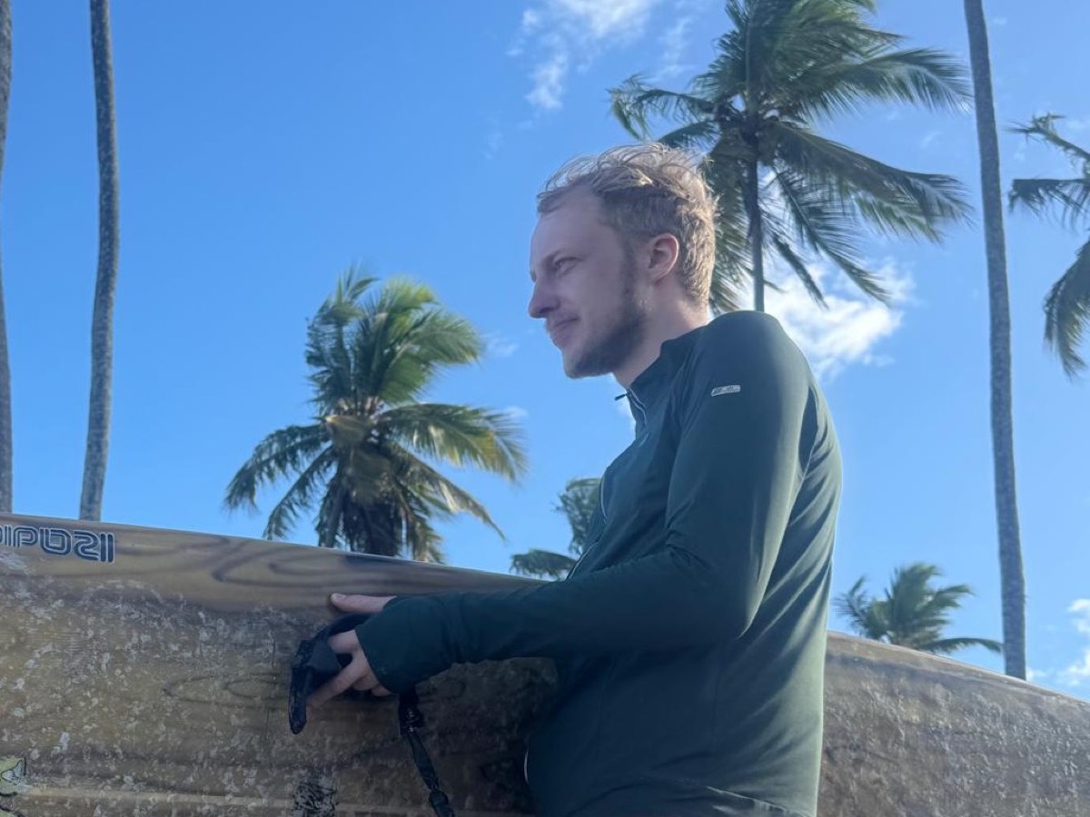 Johannes steht mit einem Surfbrett vor Palmen und blauem Himmel