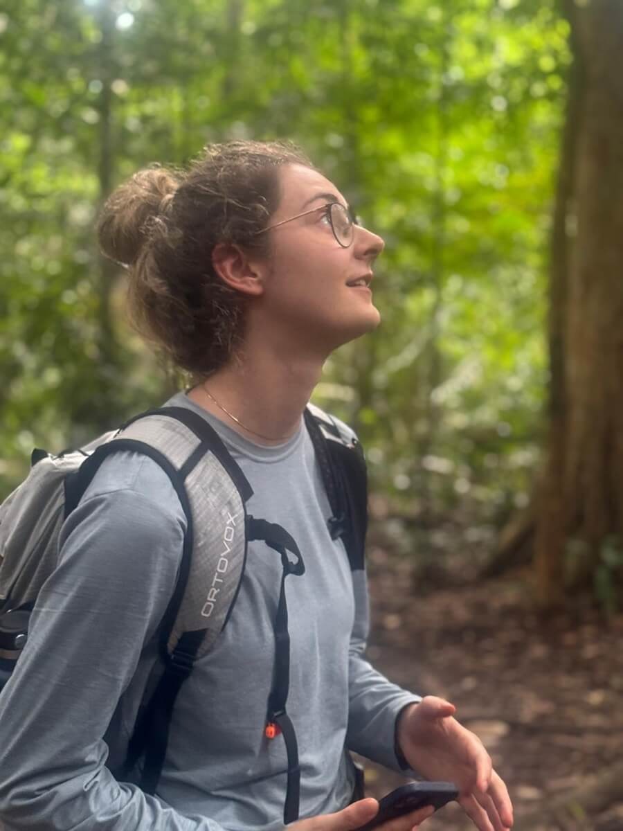 Selina steht mit Rucksack im Regenwald und blickt nach oben in die Baumkronen